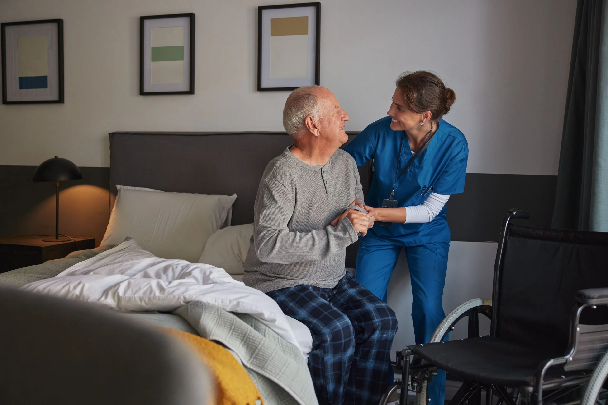 A caregiver helping an older man move safely beside his bed during recovery